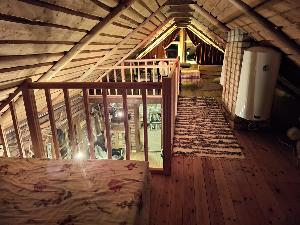 An open loft area reveals wooden beams and a sloped ceiling. A cozy bed with floral bedding is positioned near a wooden railing. Soft lighting highlights a plush rug on the floor, while a subtle view of the lower level is visible through the railing.