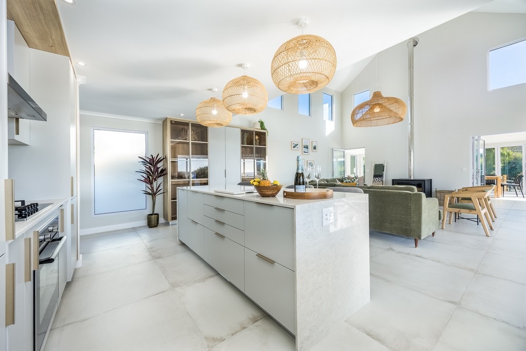 A modern kitchen is showcased, featuring a central island with storage and a light-colored stone countertop. Natural light filters through large windows. Pendant lights hang from the ceiling above, complemented by cabinetry and an open space connecting to the living area.