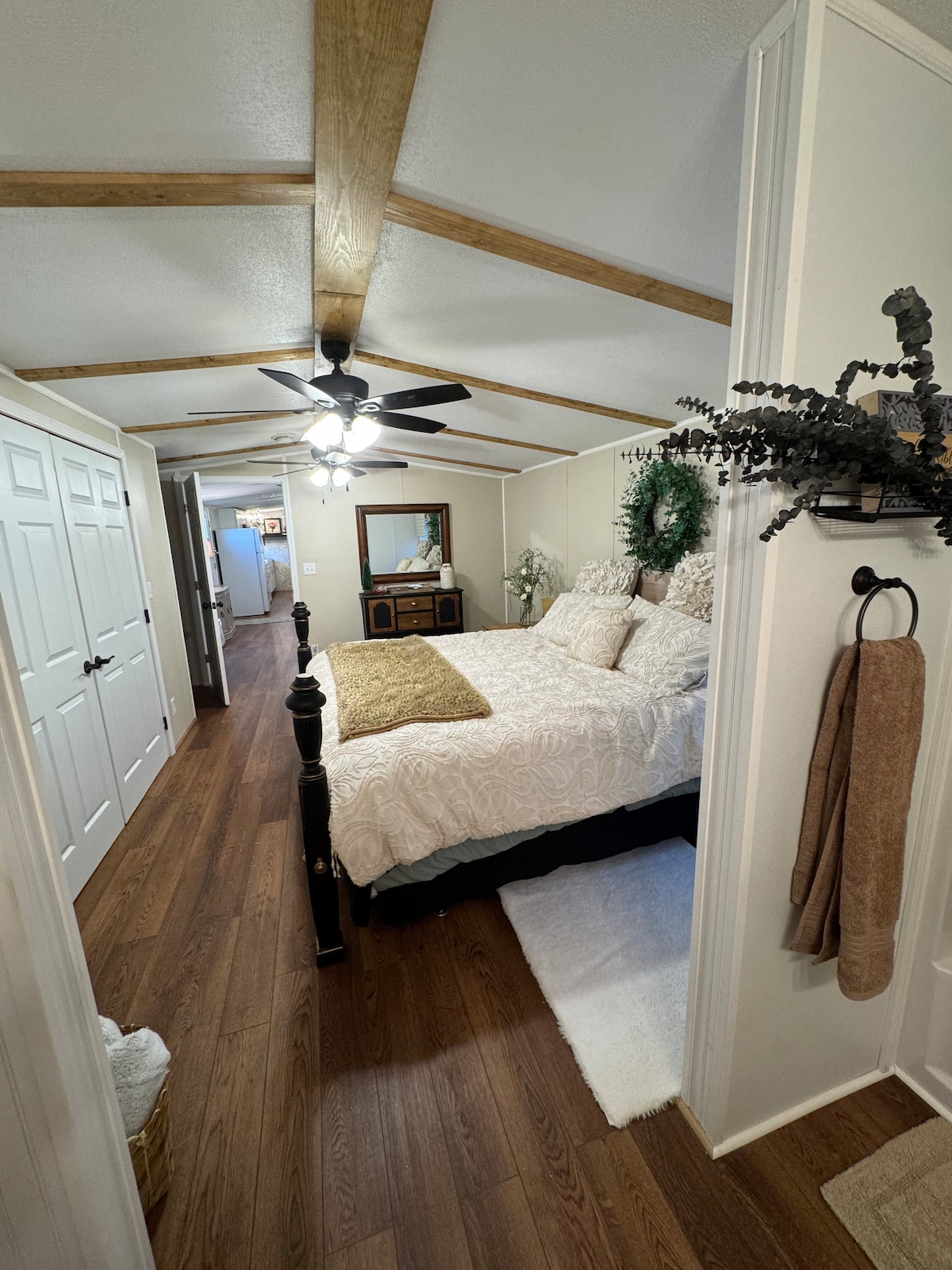 A spacious bedroom is featured, showcasing a dark wood bed frame with a soft white comforter and matching accent pillows. Light hardwood flooring contrasts with the room's neutral walls. A ceiling fan is visible above, and a decorative mirror and greenery adorn the dresser, enhancing the tranquil space.