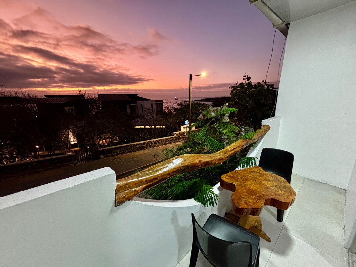 A private balcony is displayed, featuring a natural wood table surrounded by a black chair. Green foliage is seen alongside the table, and a soft sunset sky with pink and purple hues is visible in the background. Street lights illuminate the area gently.