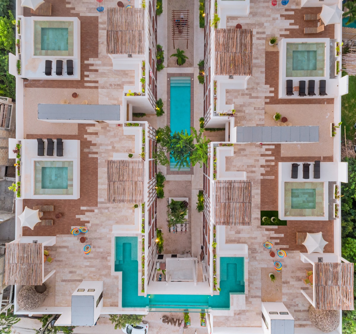 An aerial view of the condominium complex reveals a symmetrical layout featuring multiple pools and lounge areas. Sun loungers are positioned along the sides of the pools, surrounded by lush greenery and elegant landscaping. The overall design reflects a harmonious blend of nature and luxury.