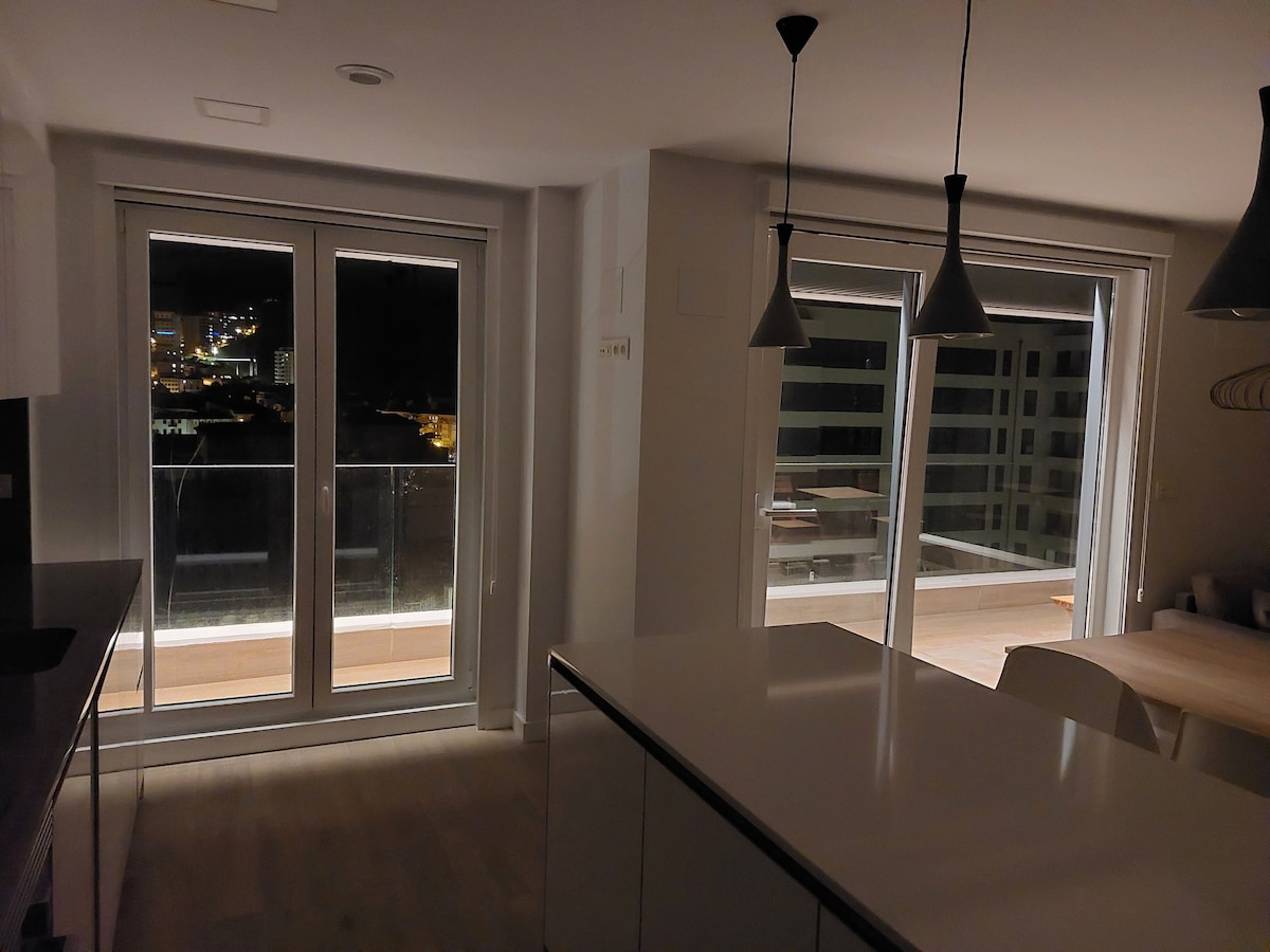 A modern kitchen area is visible, featuring a large island countertop and sleek cabinetry. Floor-to-ceiling glass doors open to a spacious balcony, allowing for natural light. Night views of the city skyline can be seen partially illuminated through the glass.