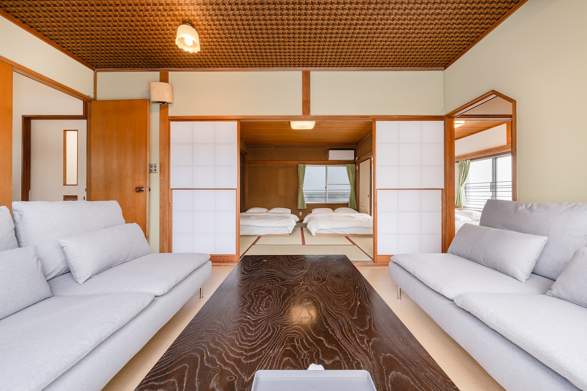 A spacious living area is presented with two light gray sofas facing a dark wooden table. Sliding shoji doors separate the room from two sleeping areas, which feature futons laid out on the tatami mat. Natural light filters in, creating a serene atmosphere.