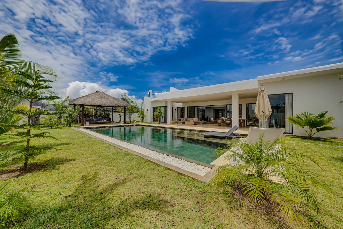 The villa features a modern silhouette with expansive glass doors leading to a lush green yard. A sunlit terrace borders a heated infinity pool, complemented by a shaded gazebo, ideal for relaxation. Palm trees and tropical foliage add to the serene outdoor setting.
