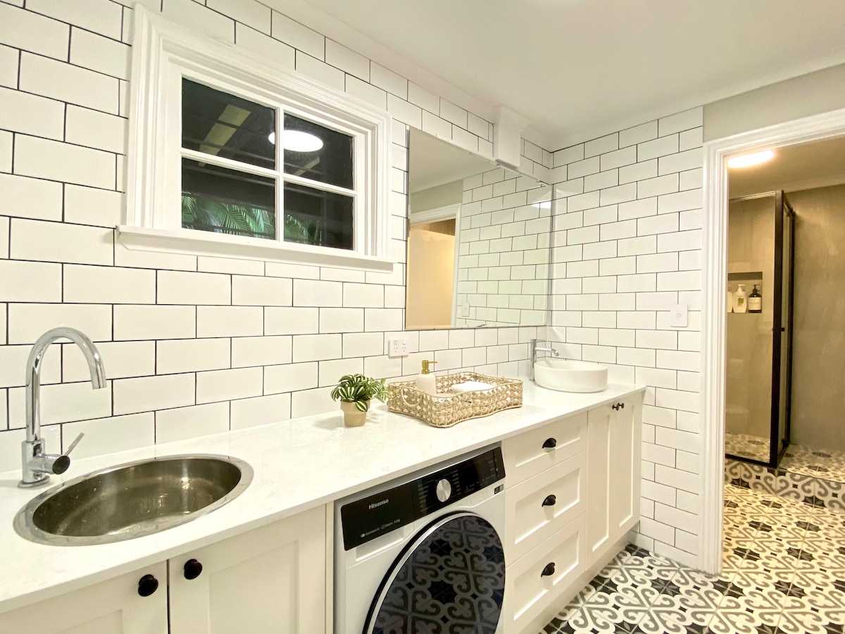 The bathroom features white subway tile walls, a spacious vanity with an integrated sink, and a washer and dryer beneath. A large mirror reflects the fresh design, while a decorative plant adds a touch of greenery. A glass shower enclosure is visible in the background.