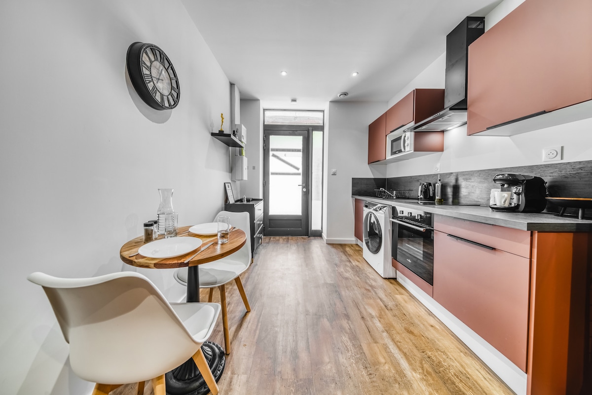 A modern kitchen space is highlighted by sleek cabinetry in warm tones. A small round dining table with two chairs is set for meals, positioned near a door that leads outside. Essential appliances, including a washer-dryer, microwave, and coffee maker, are integrated seamlessly along the countertops.
