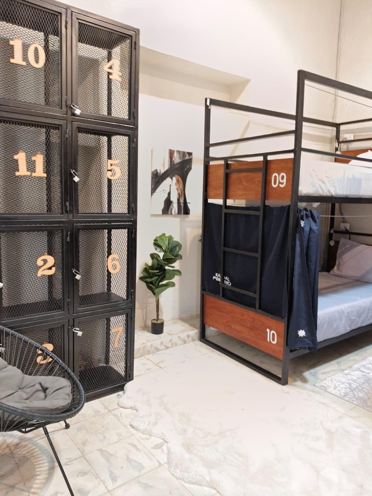 A modern interior is showcased with a metallic bunk bed and individual storage lockers featuring numbers. A small potted plant is positioned near a low chair, and a wall painting adds a subtle touch of decor. Soft rugs cover the floor.