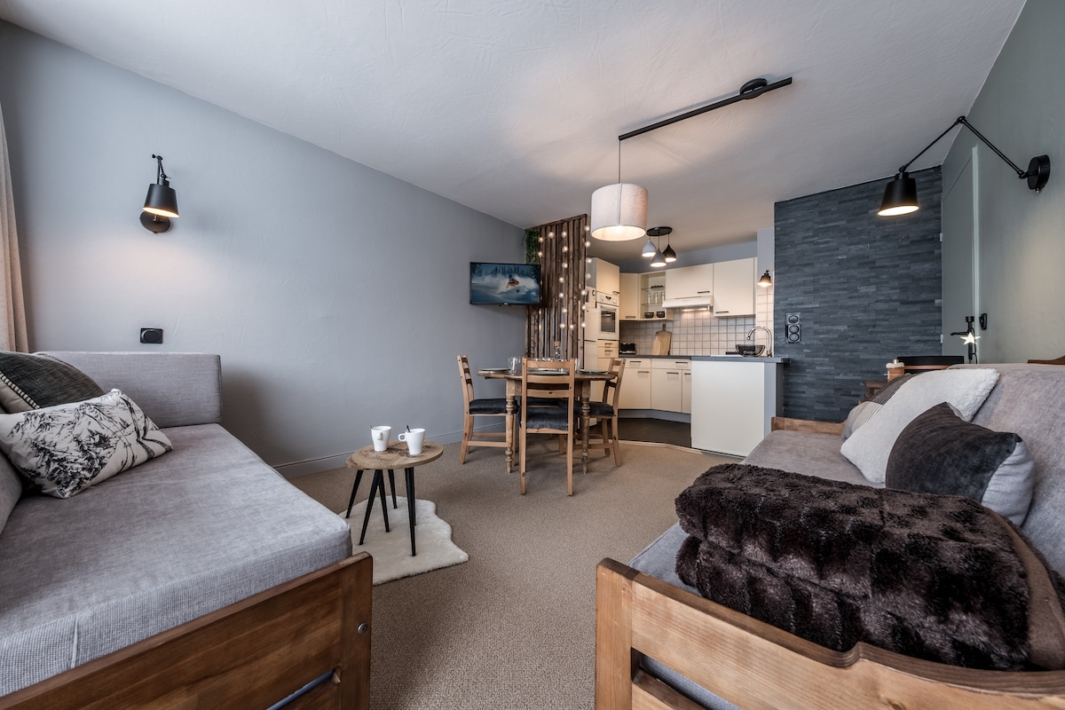 A comfortable living area features two sofa beds with plush throws, arranged around a wooden coffee table. A dining area with wooden chairs is visible, leading to a well-equipped kitchen with modern appliances. Natural light fills the space, complemented by stylish wall sconces.