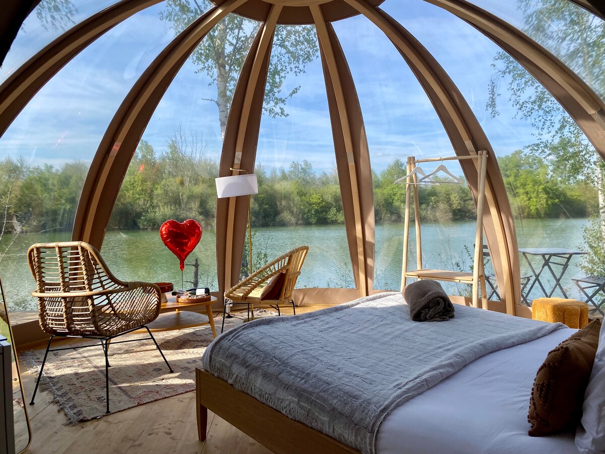 An inviting sleeping area features a spacious bed with a cozy blanket, surrounded by a unique dome structure. Two rattan chairs provide comfortable seating, alongside a small table. Large windows offer a view of the serene water and lush greenery just beyond.