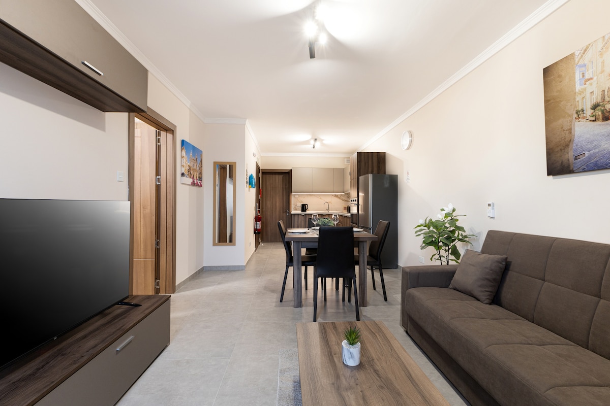 A spacious living area is presented with a neutral color palette. A comfortable sofa faces a large flat-screen TV, while a dining table is visible in the fully equipped kitchen in the background. Soft lighting enhances the overall ambiance, creating a welcoming environment.