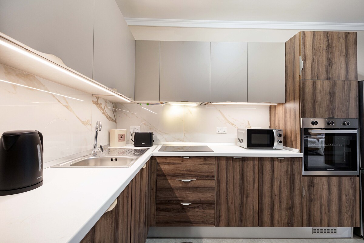 The kitchen features a modern design with wooden cabinets and sleek countertops. Essential appliances include a kettle, microwave, and oven, all integrated into the cabinetry. Under-cabinet lighting enhances visibility, while the spacious layout allows for easy movement during meal preparation.
