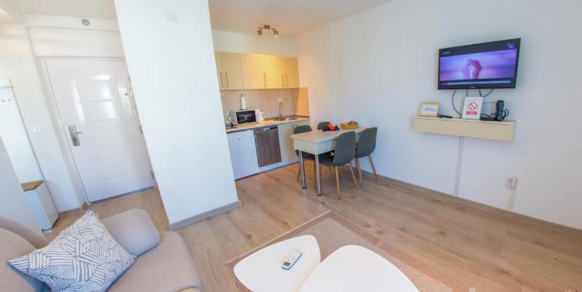 The image showcases a modern kitchen area adjacent to a cozy living space. A dining table with four chairs is visible, accompanied by a kitchen countertop with appliances. A wall-mounted TV is present, while natural light brightens the light-colored wooden flooring.