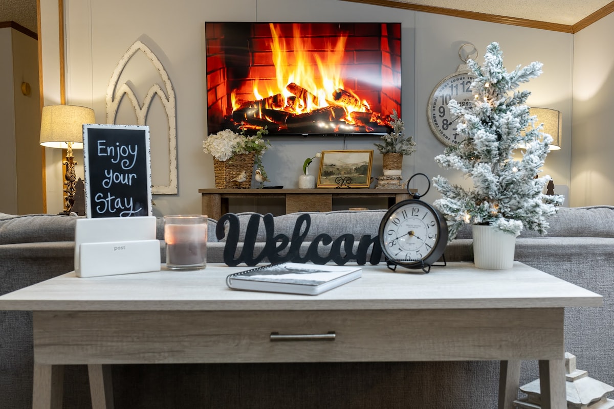 A warm living area is highlighted by a wall-mounted TV displaying a cozy fireplace. A wooden table features a welcome sign, decorative clock, a candle, and a small potted tree. Soft lighting is provided by lamps on either side of the room.