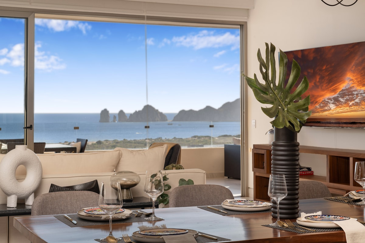 The dining area is adorned with a modern wooden table set for four, featuring elegant dishware. A large window offers a stunning view of the ocean and distant Land's End. A decorative plant and stylish accents complement the inviting space.