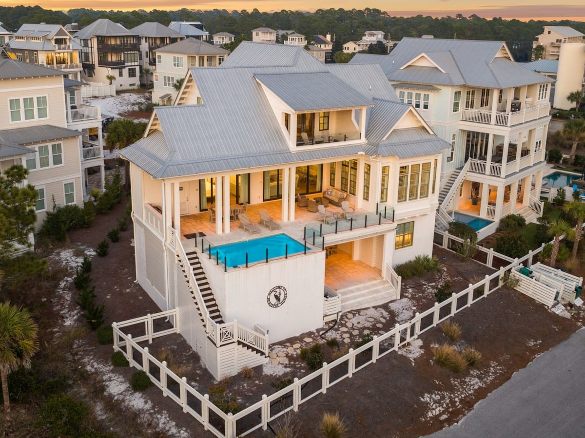 An aerial view of a spacious two-story beach house, featuring a private pool on the second floor. The structure is surrounded by well-maintained landscaping, with an adjacent wooden fence. Bright exterior walls complement a gray metal roof, providing a stylish contrast to the natural surroundings.