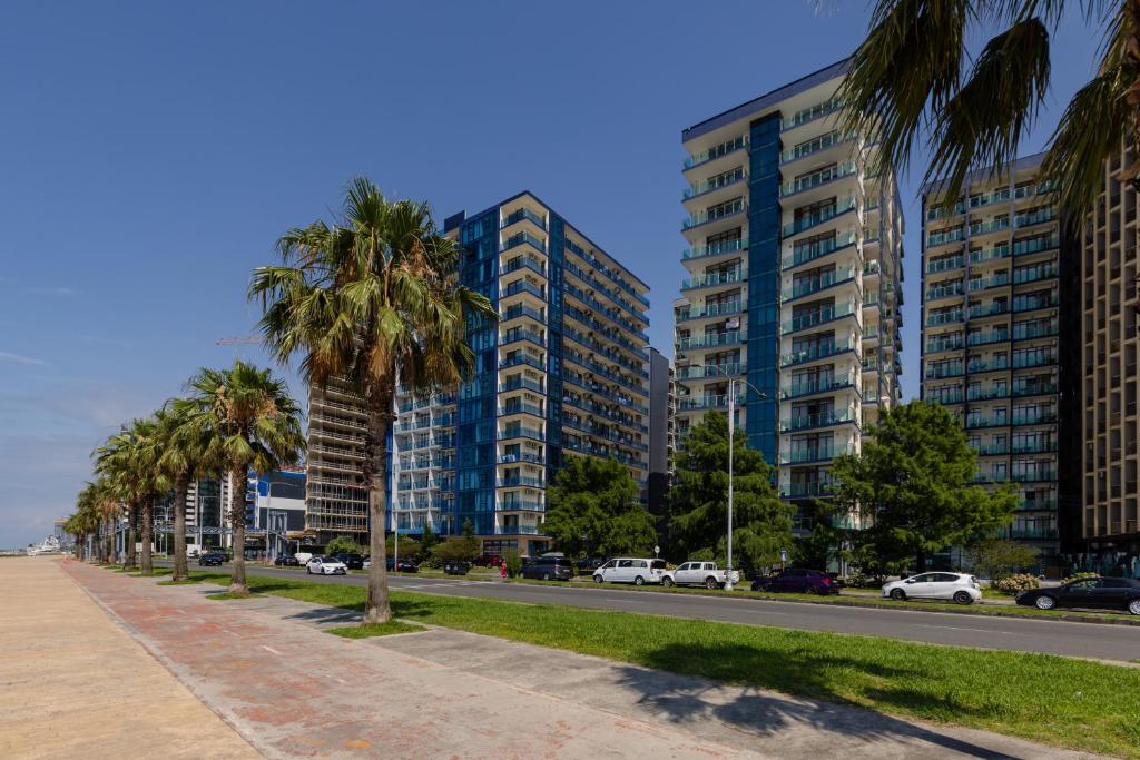 A row of modern high-rise buildings is flanked by tall palm trees along a spacious walkway. The architecture features glass balconies and vibrant blue accents, while a clean sidewalk runs parallel to the roadway, providing a clear view of the surrounding landscape.