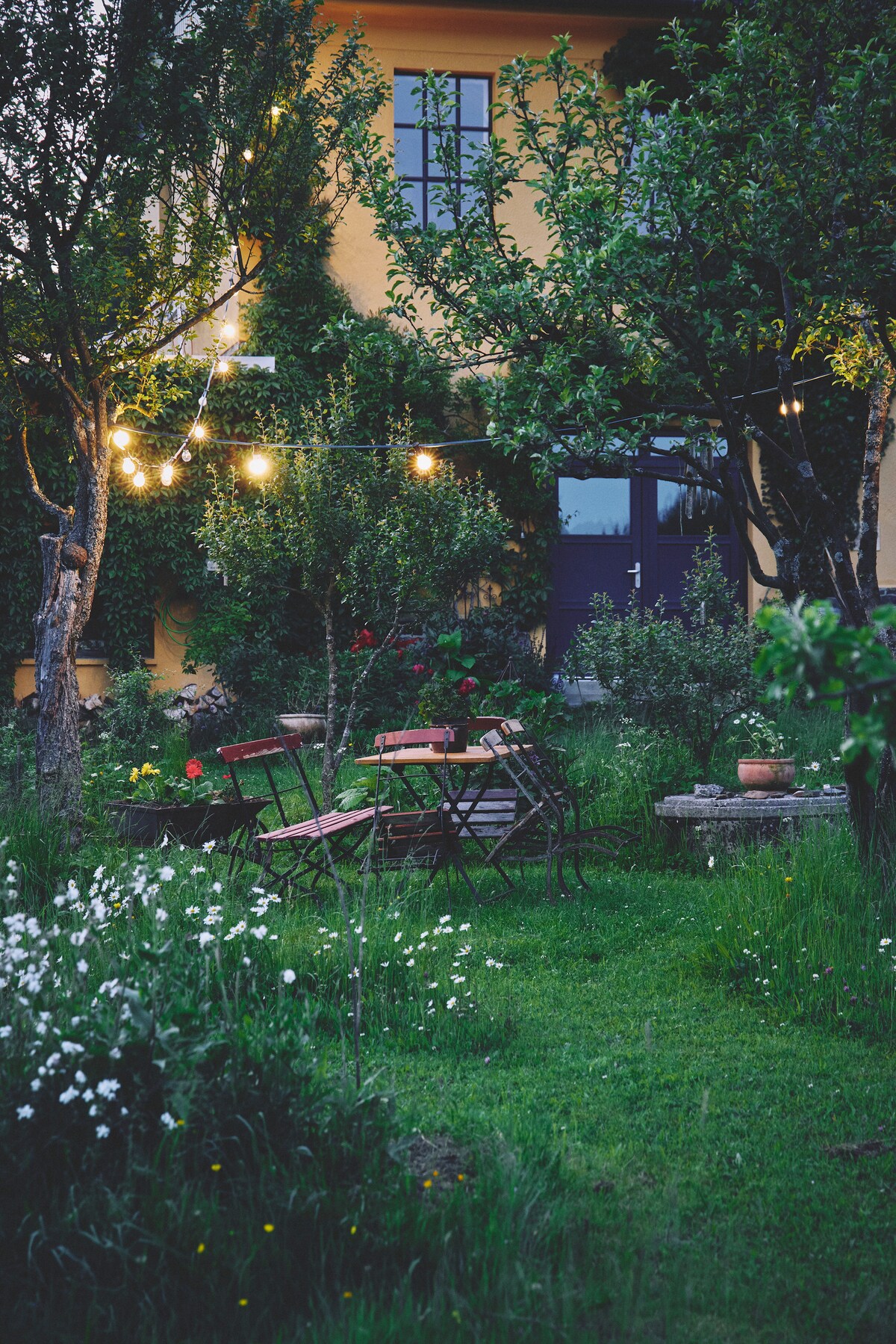 A garden area is framed by trees, featuring wooden furniture including a dining table and chairs. Soft lighting from decorative strings illuminates the space, creating a serene atmosphere. Various plants and flowers enhance the natural beauty surrounding the outdoor seating area.