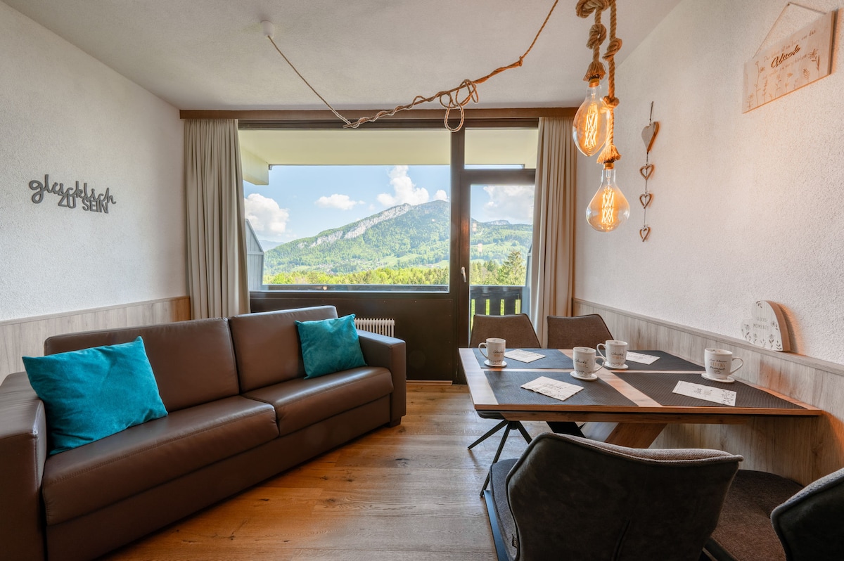 A comfortable living area features a brown sofa adorned with teal cushions, complemented by a wooden dining table set for four. Large windows frame a scenic mountain view, while warm pendant lights hang above, creating a cozy ambiance.
