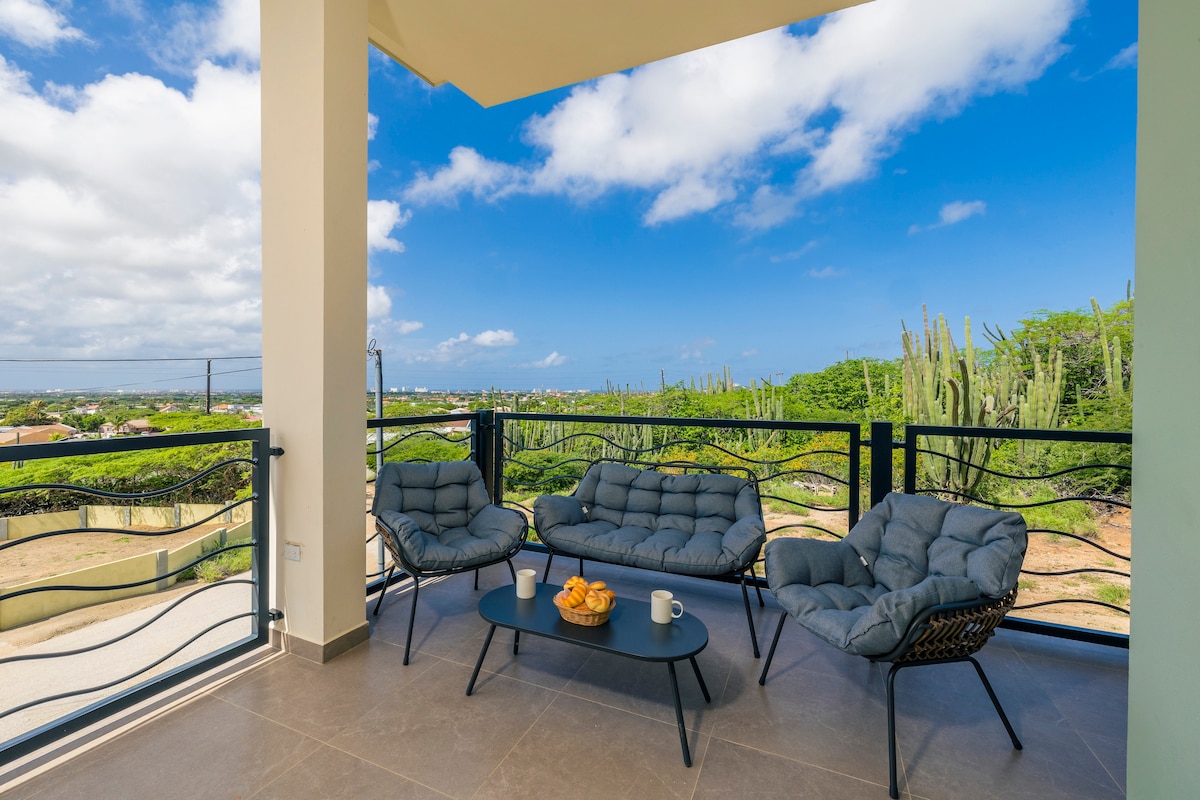 A covered balcony space is furnished with comfortable seating and a small coffee table. The surrounding scenery includes lush greenery and views of the landscape, with a bright blue sky and fluffy white clouds overhead.