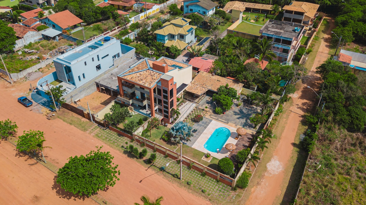 An aerial view captures a spacious property featuring a private pool, surrounded by landscaped gardens and sun loungers. The nearby structures showcase varied architectural styles. A sandy road leads to the home, suggesting easy access to the area.