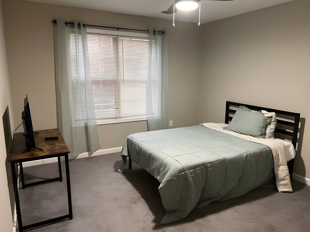 غرفة نوم واسعة بسرير فاخر، مزينة بوسائد زرقاء وأعمال فنية تجريدية على الحائط. تؤدي الغرفة إلى مدخل مع سلالم، مما يضيف عمقًا إلى المساحة الساطعة والبسيطة.