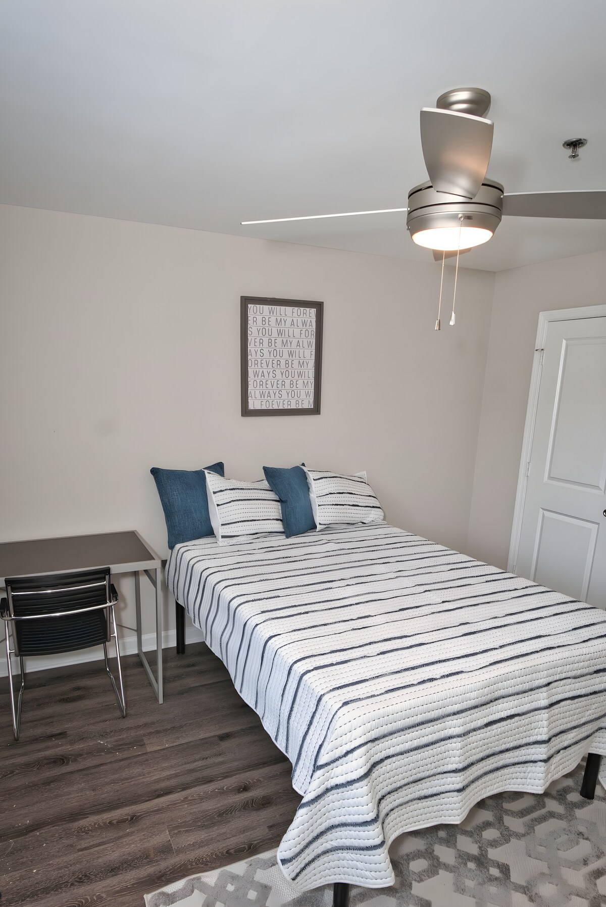 غرفة نوم بسيطة تحتوي على سرير مع لحاف أخضر فاتح ووسادة متطابقة، موضوعة مقابل جدار بيج محايد. نافذة مع ستائر خضراء شفافة تسمح بدخول الضوء الطبيعي الناعم. 