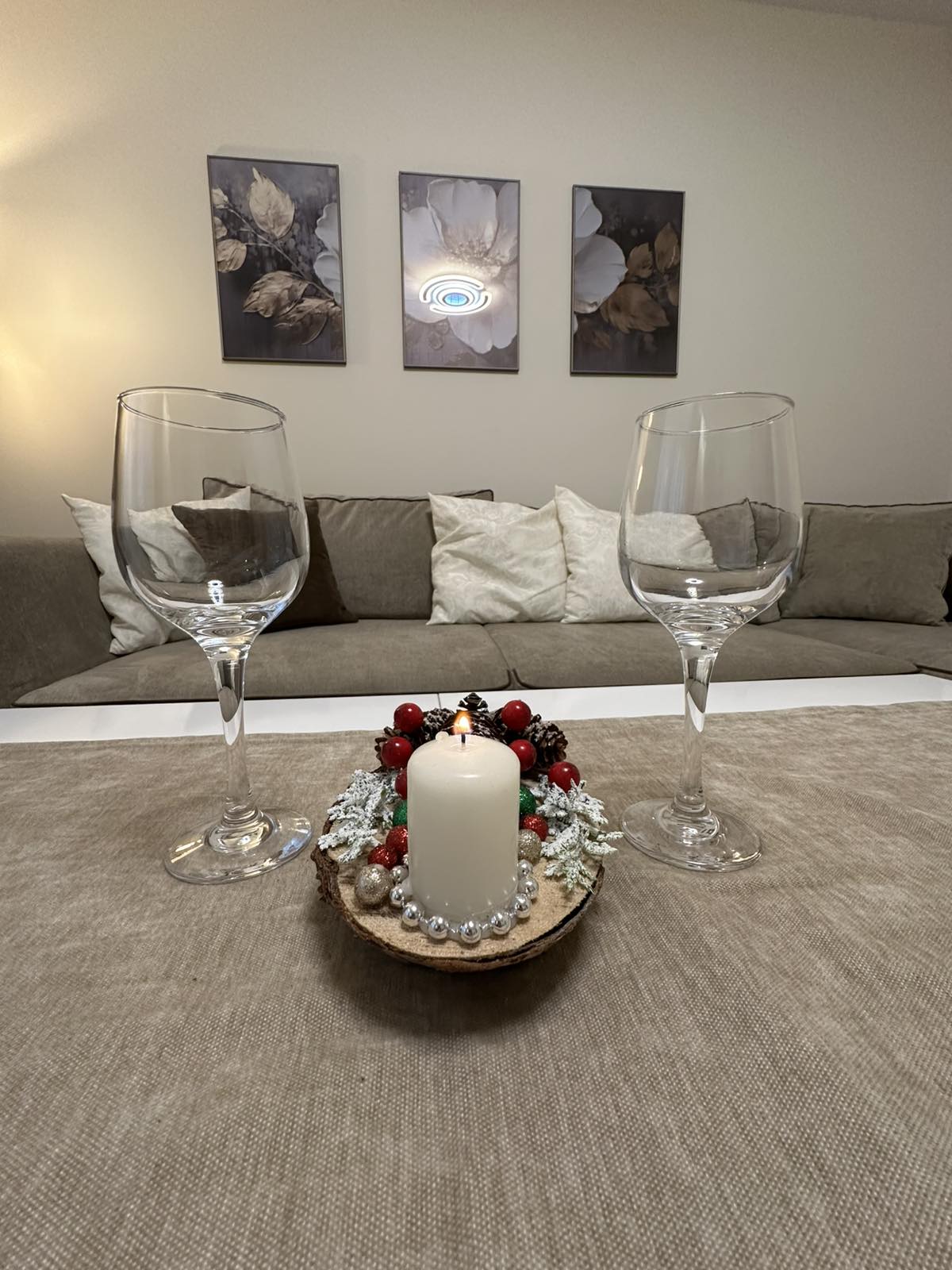 Two wine glasses are positioned on a neutral-toned table. In the center, a decorative arrangement features a candle surrounded by festive elements such as red berries and artificial snow. A cozy sofa adorned with various throw pillows is visible in the background.