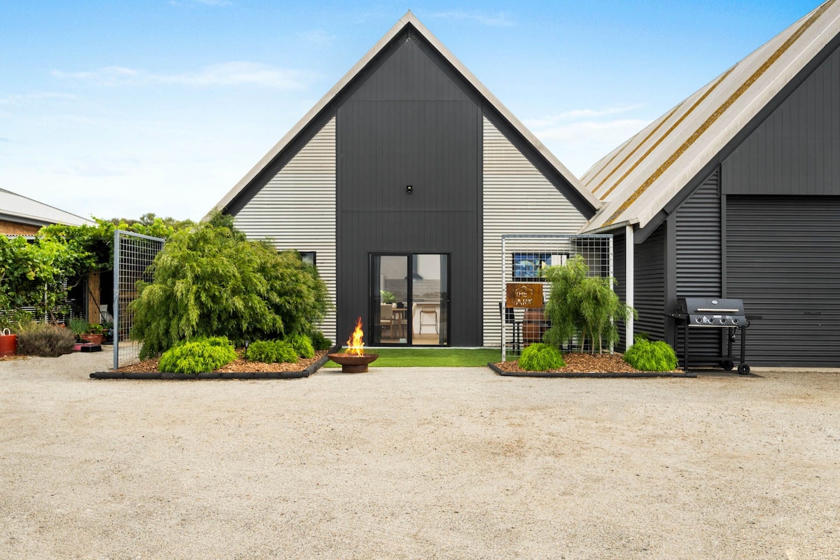 A striking barn conversion exterior is displayed, featuring a combination of sleek black and silver panels. Lush greenery surrounds the entrance, with a small fire pit set in a gravel area, and a BBQ station is positioned nearby on the right.