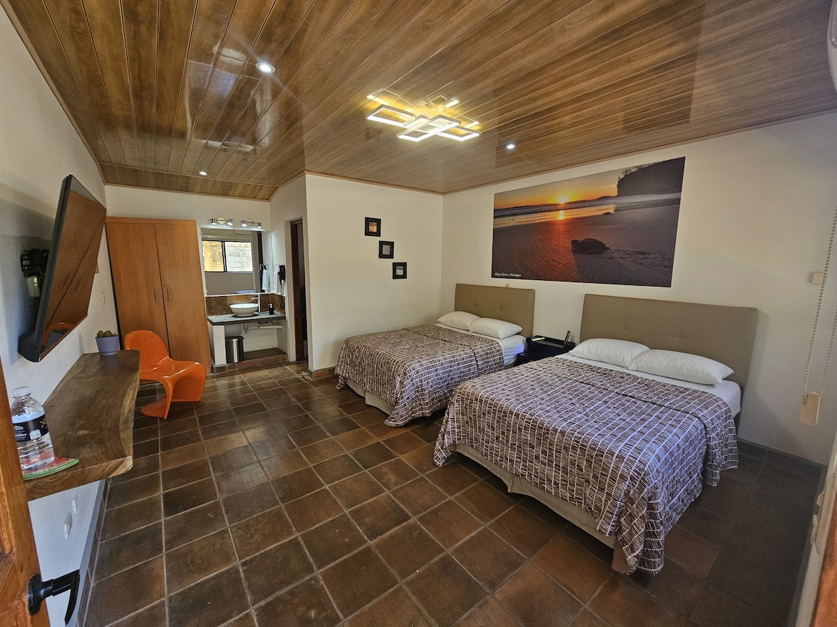 The room features two spacious beds with neutral-colored linens. A wooden ceiling and tiled floor add warmth. A vibrant sunset photograph adorns the wall, while a television is mounted. A small seating area with an orange chair complements the natural light from windows adjacent to the bathroom.