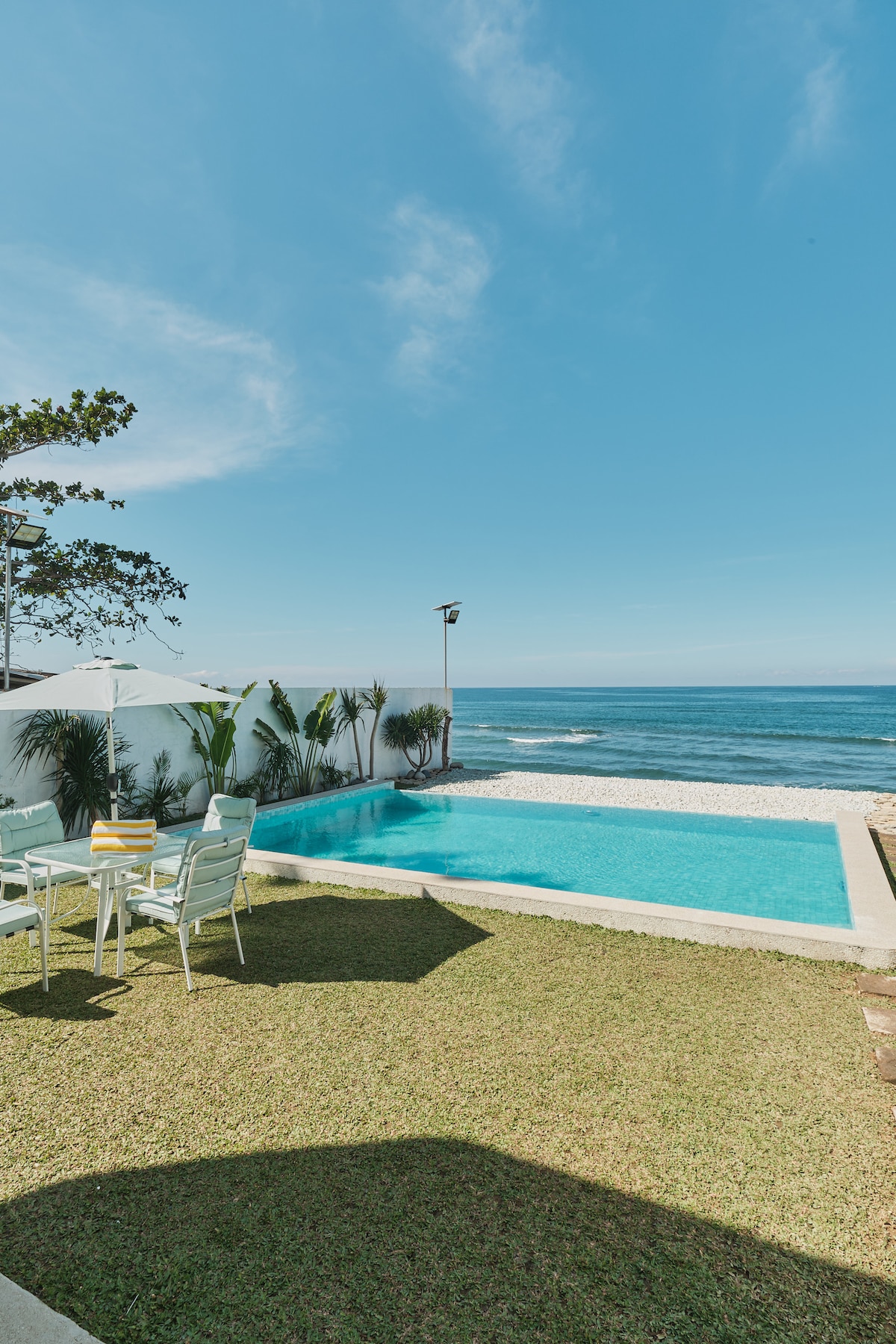 A private swimming pool is surrounded by a well-maintained grassy area, offering views of the ocean. A small dining set with light-colored chairs is positioned nearby, providing a space for outdoor meals. Clear skies and gentle waves are visible in the background.