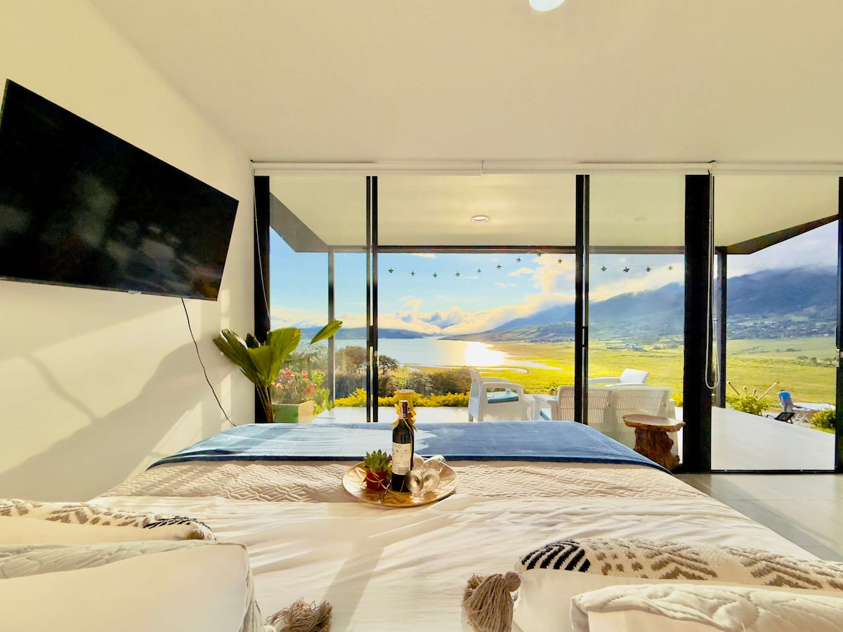 A spacious bedroom features large glass doors that open to stunning views of the lake and mountains. The area includes a large bed with decorative pillows and a small tray with a bottle of wine and snacks. Natural light fills the room, enhancing the serene atmosphere.