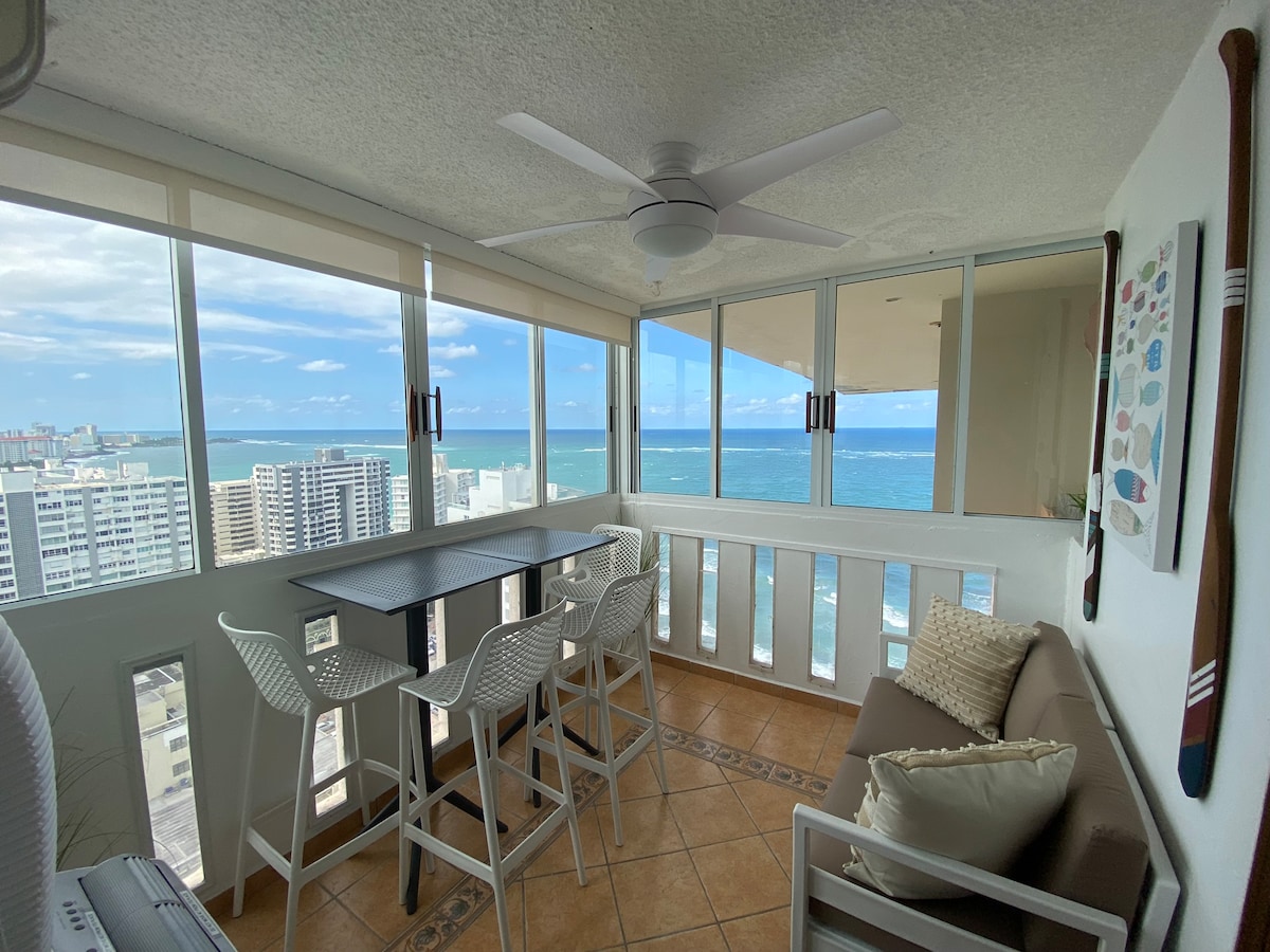 An enclosed balcony provides expansive ocean views through large glass windows. A modern table with high stools is positioned next to a comfortable sofa. Neutral tones are complemented by coastal-themed decor, creating a serene environment for relaxation and enjoyment of the surroundings.