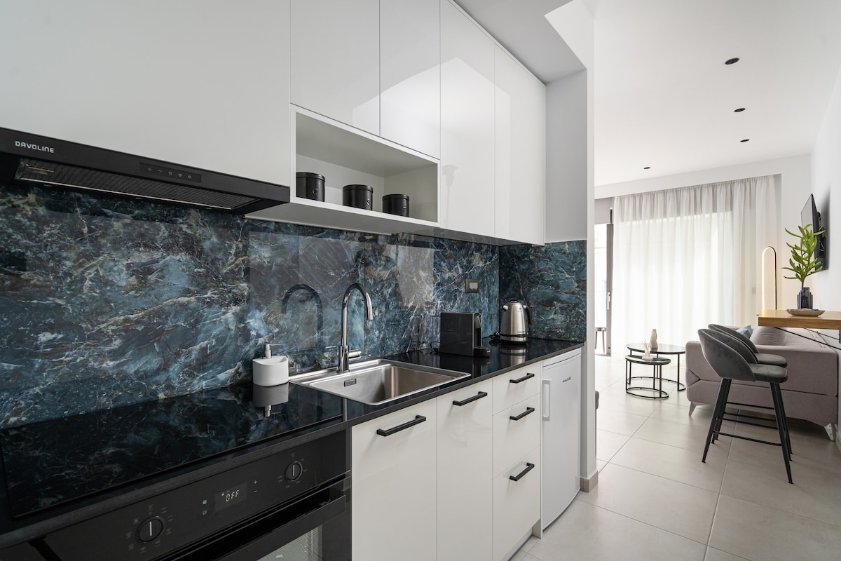 The modern kitchen features sleek white cabinetry complemented by a striking blue marble backsplash. Stainless steel appliances include an electric oven and kettle. The open-plan design allows for natural light to brighten the space, with a cozy seating area visible in the background.