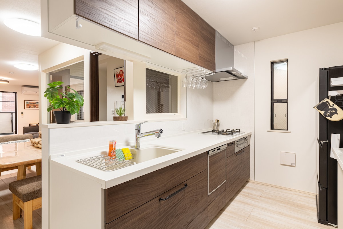 A modern kitchen features sleek wood cabinetry and a spacious countertop. Essential appliances, including a stove and refrigerator, are neatly arranged. Natural light filters through a small window, enhancing the clean and functional layout, complemented by a potted plant on the counter.