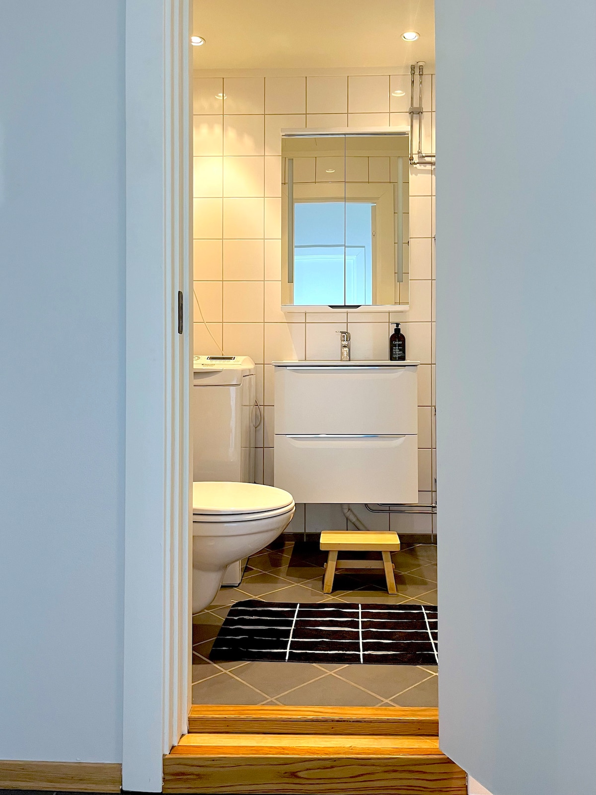 A well-lit bathroom is visible through an open door, featuring white tiled walls and a modern sink with a mirror above. A small stool is placed in front of the toilet. A black and white rug adds texture to the tiled floor.
