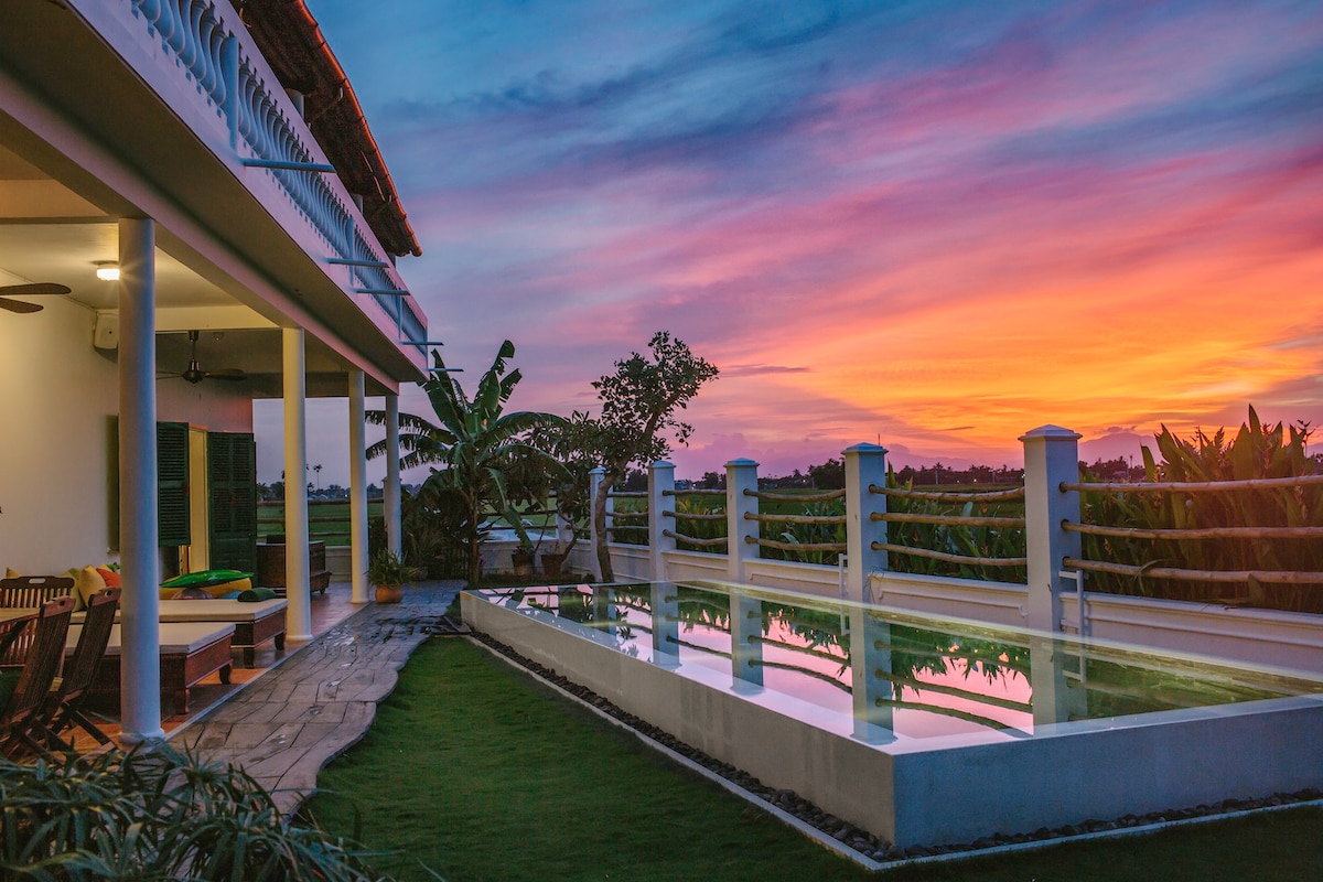 The outdoor area features a modern swimming pool reflecting a vibrant sunset, bordered by lush greenery. A spacious terrace with comfortable seating invites relaxation, while the evening sky presents a gradient of warm colors, enhancing the peaceful ambiance of the surroundings.