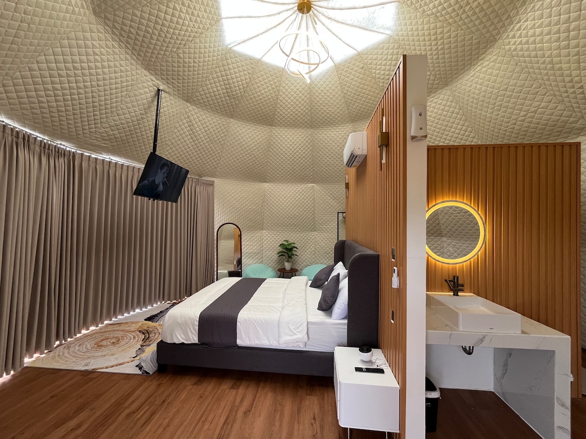 A modern bedroom features a spacious bed with neutral linens, complemented by a circular mirror and decorative items. Warm wooden accents are present, alongside a unique geometric ceiling design. Natural light filters through the curtains, enhancing the overall ambiance of the space.