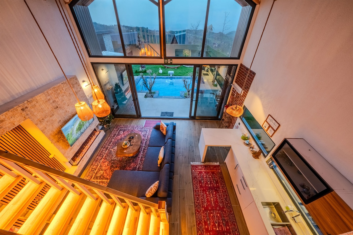 An aerial view of the open living space showcases a cozy corner sofa with decorative cushions, warm wooden flooring, and a rug. Large glass doors provide a view of the private garden and pool, while a modern kitchen area is visible in the background.