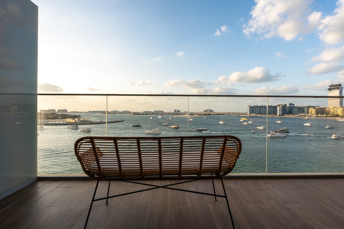 A balcony overlooks the water, with a woven lounge chair positioned to enjoy the view. The horizon features boats navigating the bay, and modern buildings are visible in the distance. Soft clouds drift across a blue sky, adding to the serene atmosphere.