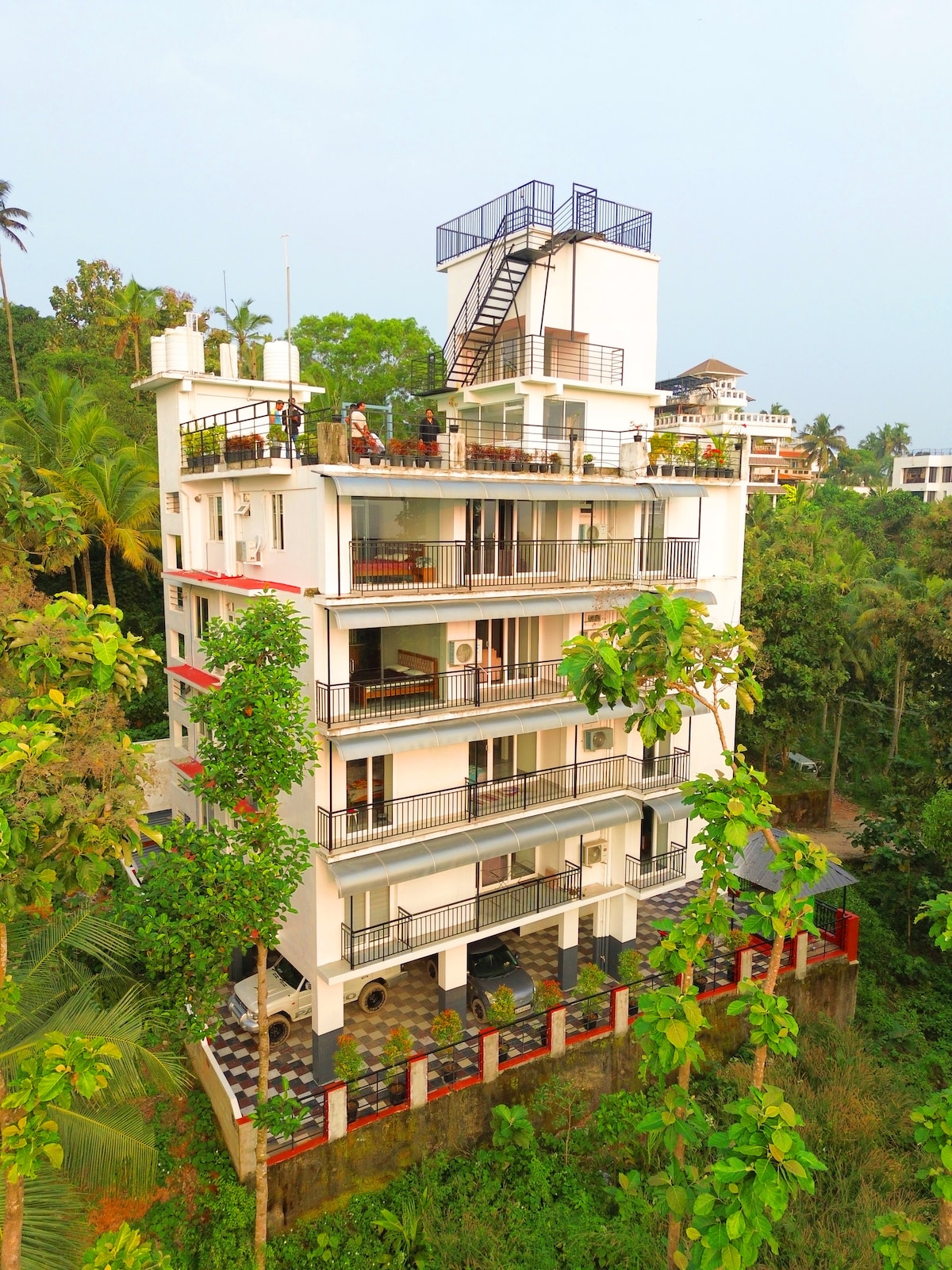 The multi-story building is set among lush greenery, with balconies on each floor offering views of the surrounding landscape. A stairway can be seen leading to the rooftop terrace, complementing the modern architectural design of the structure.