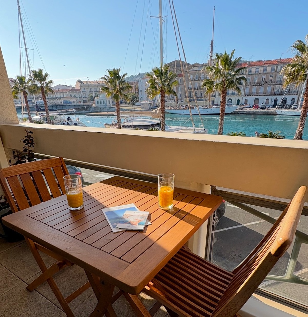 A cozy balcony scene features a wooden table set for two, adorned with two glasses of orange juice. The view showcases palm trees lining the promenade and a serene canal with sailboats and yachts visible in the background.