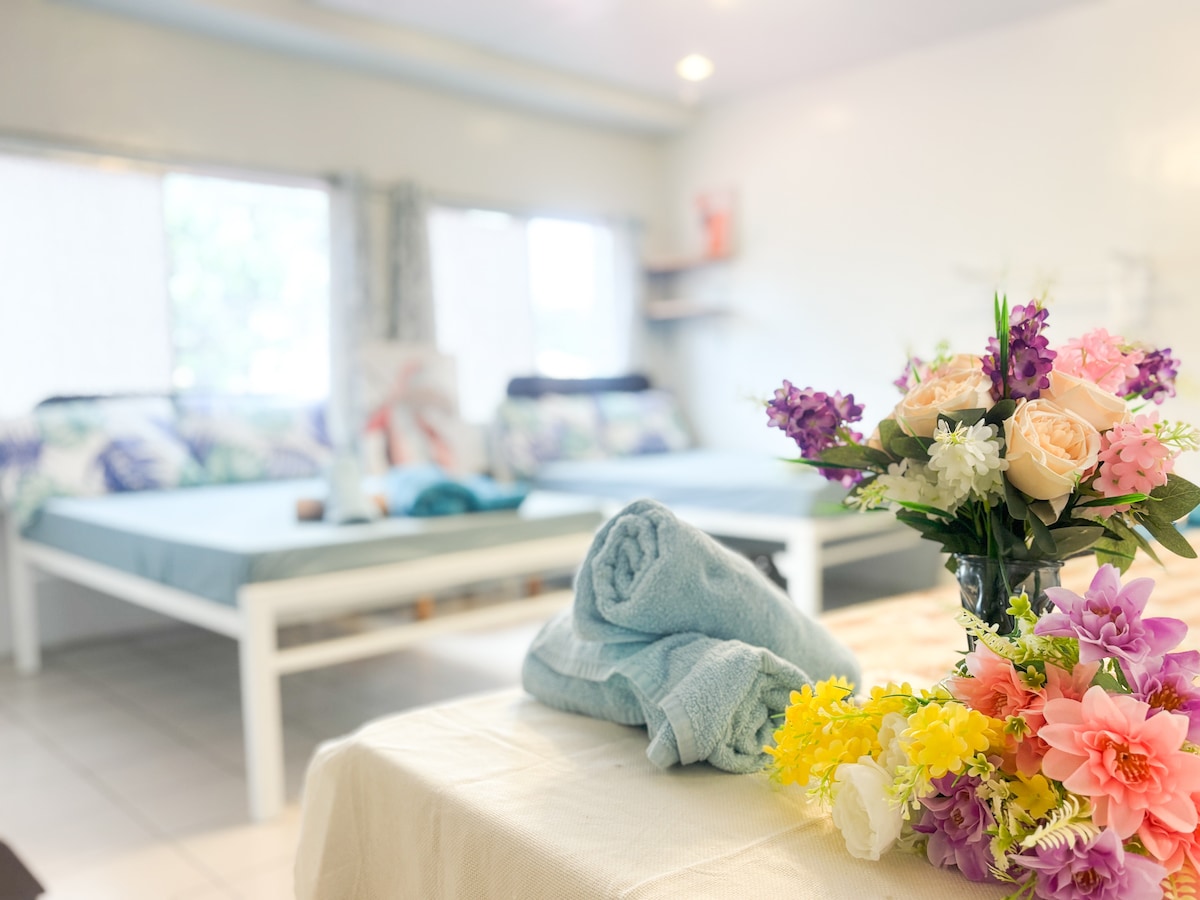 A spacious room is observed, featuring two beds adorned with decorative pillows. In the foreground, a table with a soft towel and a vibrant bouquet of flowers adds a touch of color. Natural light filters in through large windows dressed in light curtains.