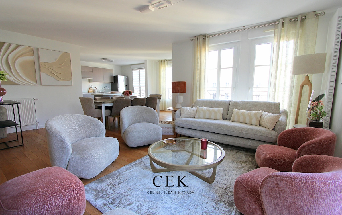 A spacious living area is furnished with soft, curved chairs in muted tones and a light sofa. Natural light fills the room through large windows, complemented by sheer curtains. A round coffee table sits at the center, resting on a patterned area rug.