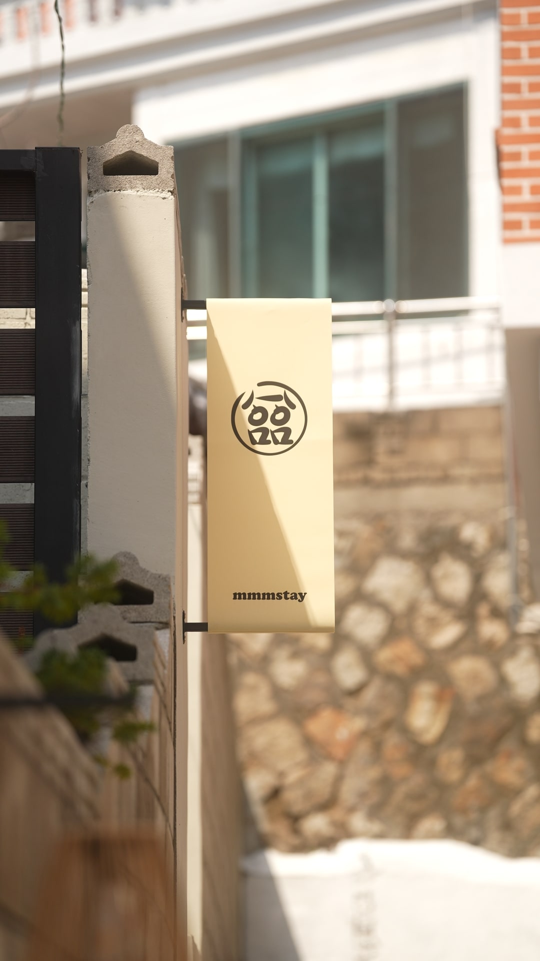 A beige banner is displayed prominently, featuring a simple logo representing the property. The banner is fixed securely to a vertical pole, with a blurred background of a residential area, showcasing brick and stone architecture.