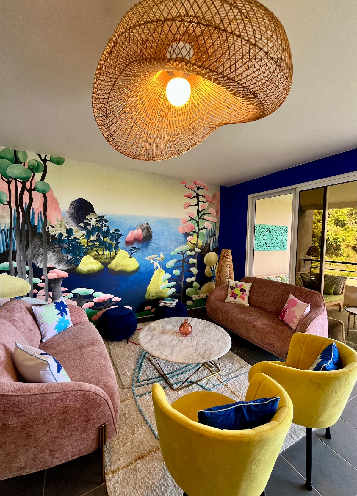 A vibrant living area features two rounded pink sofas and two yellow chairs, inviting relaxation. A circular coffee table is centered on a textured rug. A colorful mural depicting a serene landscape enhances the backdrop, while large windows provide natural light and views of the outdoors.