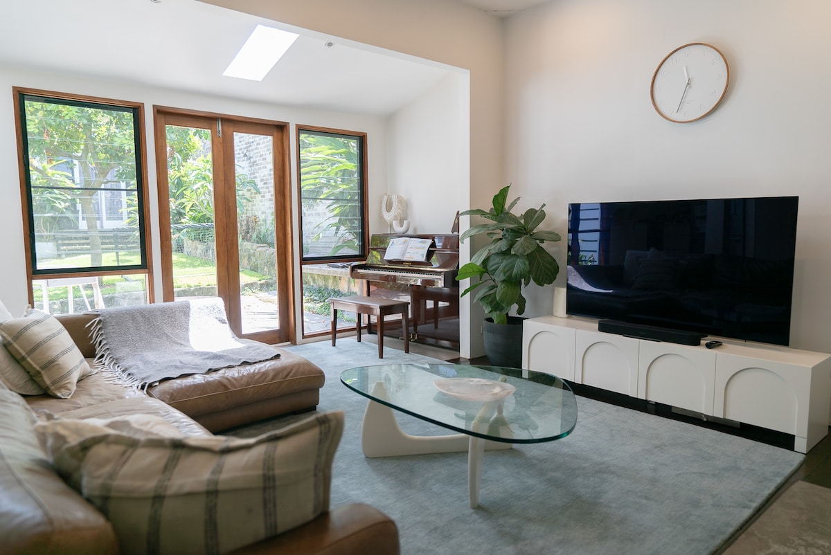 A light-filled living area features large glass doors that open to a garden view. A modern glass coffee table sits in front of a comfortable leather sectional sofa. A television and a piano are present, with a decorative plant adding a touch of greenery.
