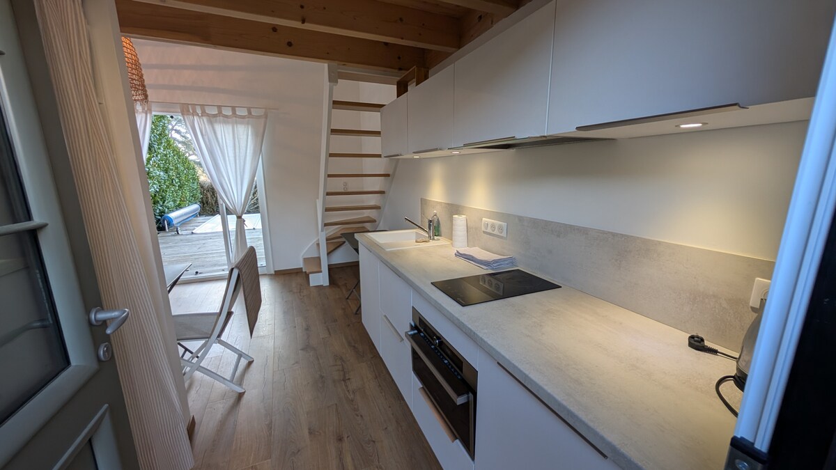 A modern kitchen showcases sleek cabinetry and a light-colored countertop. An under-counter oven and cooktop are visible, with natural light flooding in from a nearby window. A wooden staircase leads to a mezzanine, while outdoor seating is seen through an open door.