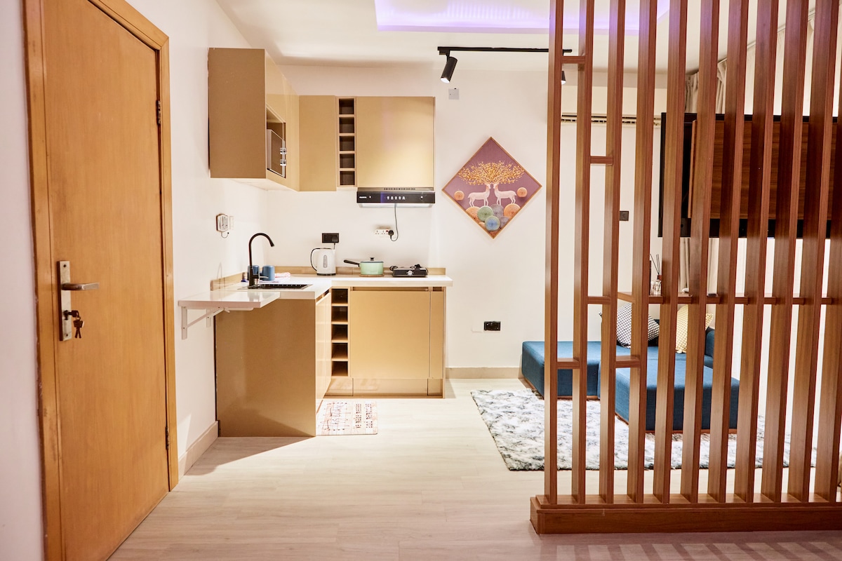 An open-plan kitchenette is depicted, featuring modern cabinetry, a microwave, a kettle, and a mini fridge. A stylish wooden partition separates the kitchen from the living area, which contains a cozy seating arrangement and a decorative piece on the wall.