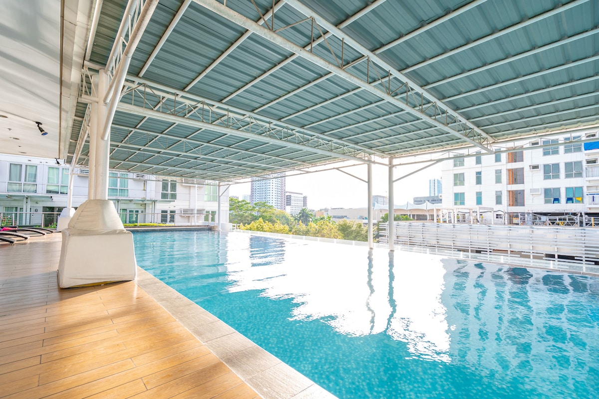 An infinity pool is showcased, featuring calm blue water that reflects the surrounding structures. Sunlight creates a shimmering effect on the surface. The pool area is complemented by wooden decking and is partially covered by a spacious overhead canopy.