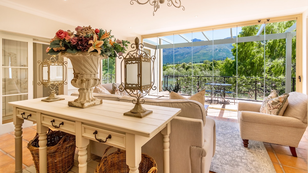 A bright and airy living space features a stylish console table adorned with decorative vases and lanterns. Large glass doors provide a view of the outdoors, framed by lush greenery and mountains, while comfortable seating invites relaxation.